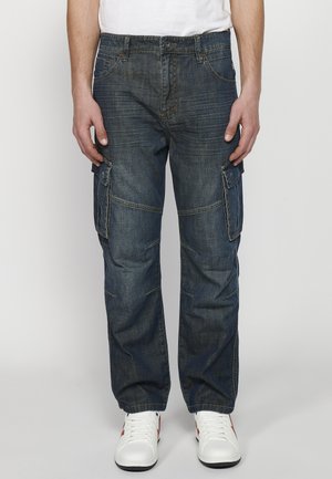 Pantaloni cargo din denim de culoare albastru închis, cu multiple buzunare laterale, croială lejeră și detalii de cusătură vizibile. Purtați cu sneakers albi.