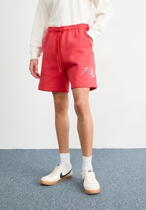 Person iført røde shorts med snøre og hvidt motiv, hvid langærmet top, hvide sokker og hvide Nike sneakers på blåt tæppe.