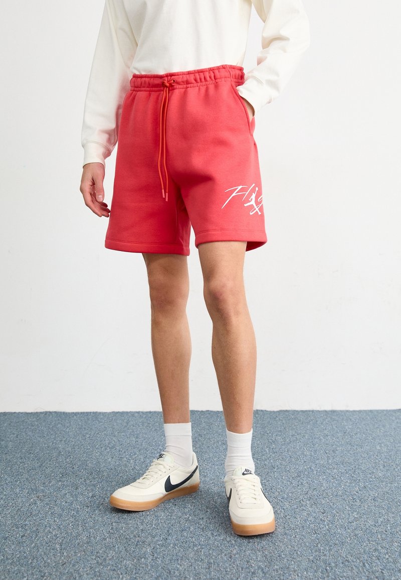 Personne portant un short rouge à cordon avec un motif blanc, un haut blanc manches longues, des chaussettes blanches et des baskets blanches Nike sur un tapis bleu.