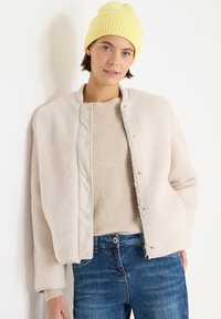 Veste bomber en polaire beige clair avec fermeture à pression, caractérisée par une texture douce, un col rond et des poignets côtelés, portée avec un pull crème et un jean bleu.
