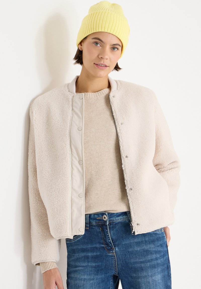Veste bomber en polaire beige clair avec fermeture à pression, caractérisée par une texture douce, un col rond et des poignets côtelés, portée avec un pull crème et un jean bleu.