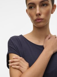 Mladá žena s krátkými tmavými vlasy v námořnickém modrém tričku, dívající se na stranu se zkříženýma rukama a jednou rukou, která se dotýká opačného ramene.