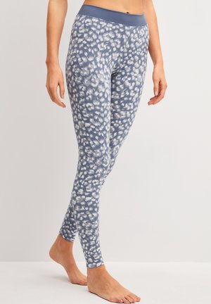 Frau trägt hoch taillierte, blau-weiße Leggings mit Punkten, steht barfuß vor einem schlichten Hintergrund.
