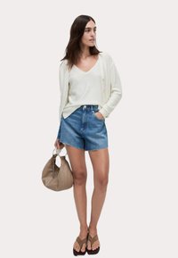 Vit, skimrande stickad kofta över en V-ringad topp, ihop med blå denimshorts, bruna flip-flops och en beige handväska.