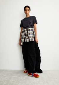 Zwarte corduroy broek, marineblauw T-shirt en een geruite flanel om de taille geknoopt. Rode en oranje sportschoenen met blauwe veters. Minimalistische achtergrond.