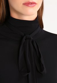 Top nero con colletto alto e dettaglio a fiocco legato al collo; texture liscia, tessuto leggero e design elegante enfatizzano la semplicità.