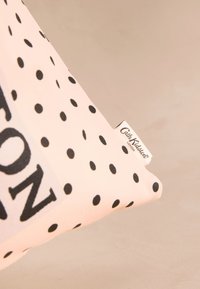 Růžový bavlněný polštář s černými puntíky a výrazným nápisem. Obsahuje malou textilní štítek s logem značky. Měkká textura a obdélníkový tvar.