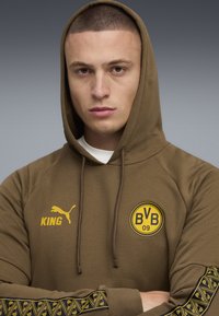 Ung mand med barberet hoved iført en olivenfarvet hættetrøje med BVB 09-logo, Puma KING-tekst i gult og dekorative striber på ærmerne, arme krydset.