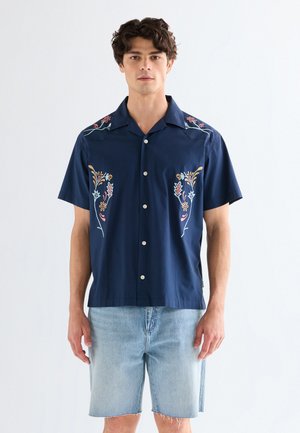 Giovane uomo che indossa una camicia blu navy a maniche corte con ricami floreali e pantaloncini di jeans azzurri, in piedi davanti a uno sfondo semplice.