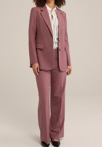 Rosa skräddarsydd kostym med en enkelknäppt kavaj och raka byxor. Matchas med en vit skjorta med svarta vertikala ränder.
