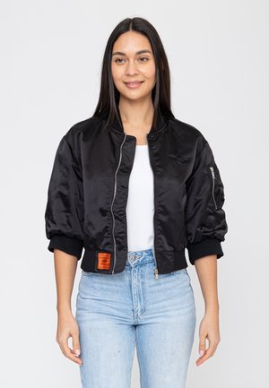 Donna che indossa una giacca bomber nera corta sopra una maglietta bianca e jeans azzurri, in piedi davanti a uno sfondo chiaro uniforme.