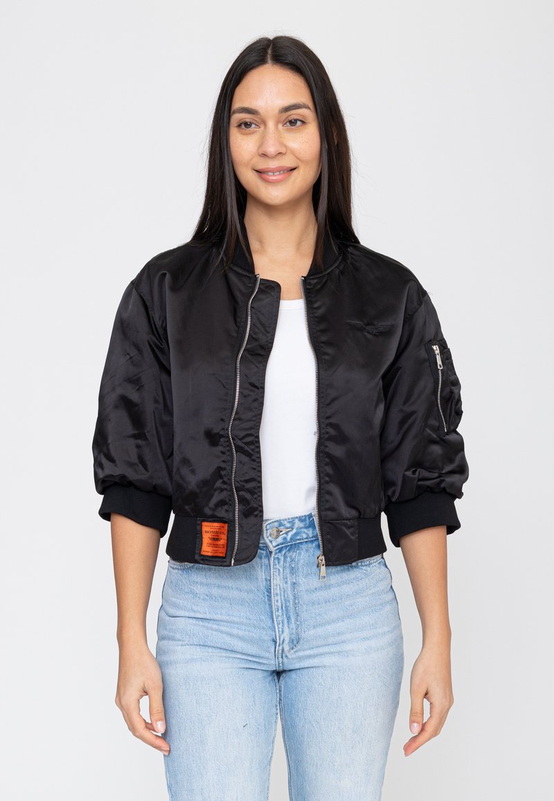 Donna che indossa una giacca bomber nera corta sopra una maglietta bianca e jeans azzurri, in piedi davanti a uno sfondo chiaro uniforme.