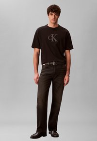 Νέος άνδρας που στέκεται, φορώντας μαύρο μπλουζάκι με το λογότυπο Calvin Klein, μαύρη ζώνη, σκούρο τζιν και μαύρα δερμάτινα παπούτσια, μπροστά σε ένα απλό γκρι φόντο.