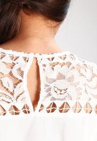 Blouse blanche avec un dos en dentelle florale, une ouverture en forme de goutte, une fermeture par boutons et des découpes géométriques complexes le long de l'encolure.