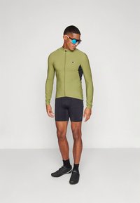 Sykkeltrøye med lange ermer i olivengrønn, med frontglidelås og svarte paneler, kombinert med svarte shorts. Modellen bærer blå solbriller og svarte sko.