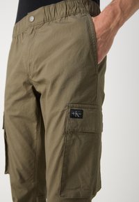 Pantalon cargo vert olive en tissu léger, doté d'une taille élastique, de poches latérales et d'une étiquette de marque sur la poche cargo.