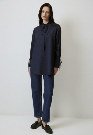 Femme portant un chemisier bleu marine foncé à manches longues avec poignets et col noués, jean droit bleu, et mocassins noirs, debout.