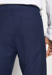 Pantaloni blu navy in tessuto testurizzato con taglio sartoriale, due tasche posteriori e passanti per cintura, abbinati a una camicia chiara.