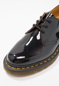 Chaussure Oxford en cuir verni noir avec bout rond, lacets plats noirs et surpiqûres jaunes le long de la semelle. Semelle extérieure en caoutchouc texturé.