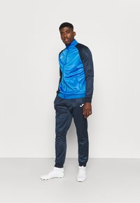 Tuta sportivo con un design blu e navy, realizzato in tessuto morbido. Include una giacca con zip e pantaloni coordinati, abbinati a scarpe da ginnastica bianche.