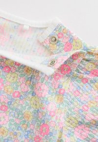 Gros plan sur un col de vêtement avec un bord blanc, tissu floral rose, jaune et bleu, avec deux boutons-pression métalliques pour la fermeture.