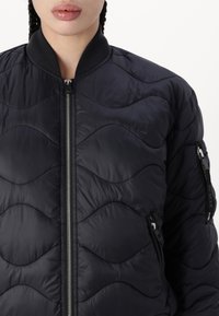 Tuvumā redzams cilvēks, kurš valkā melnu quilted bomber jaku ar sudraba rāvējslēdzi un rāvējslēdzēja kabatām uz priekšpuses un piedurknēm.