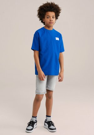 Plavi teksturirani T-shirt s kratkim rukavima s bijelom etiketom, upotpunjen svijetlosivim trapericama kratkog kroja i crnim tenisicama s bijelim detaljima.
