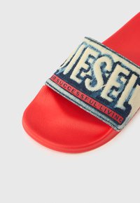 Punane kummist liugusandaal, millel on sinist teksariiet kangast rihm "DIESEL" logoga ja lisatekstiga, mis näitab tekstureeritud viimistlust.