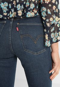 Jeans de mezclilla azul oscuro con un patrón de costura curvada en los bolsillos traseros. Presenta una etiqueta roja de Levi's. Blusa floral parcialmente visible arriba.