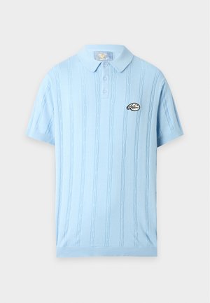 Homme grand portant un polo bleu clair à manches courtes avec logo, pantalon noir à jambes larges et baskets noires, debout sur fond blanc.