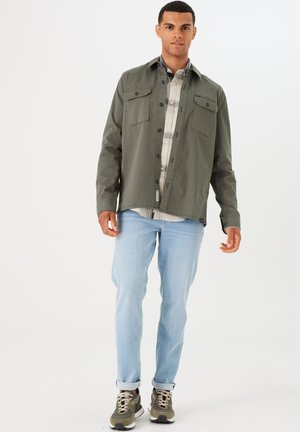 Olive green button-up jacket over a plaid shirt, light jeans, and olive sneakers. The jacket features two chest pockets and a relaxed fit.