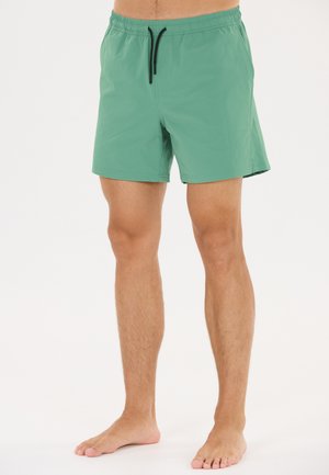 Mann trägt grüne Badeshorts mit schwarzem Kordelzug, steht barfuß vor weißem Hintergrund.
