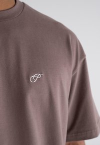 Brown cotton t-shirt featuring a crew neck and short sleeves, with a subtle white embroidered logo on the chest. Smooth texture.