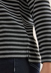 Close-up van een persoon die een zwart en grijs horizontaal gestreept gebreid trui en donkerblauwe jeans met witte stiksels draagt.