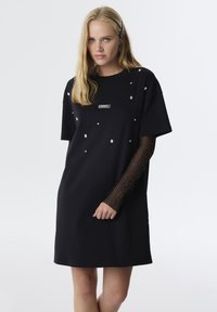 Schwarzes Kleid mit kurzen Ärmeln, das mit silbernen Verzierungen versehen ist. Transparente, strukturierte Ärmel sorgen für Kontrast. Logos sind auf der Vorderseite angebracht.