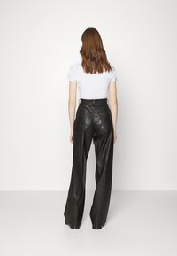 Uma mulher com cabelo castanho ondulado veste uma blusa branca de mangas curtas e calças de couro pretas de cintura alta e perna larga, de pé contra uma parede branca lisa.
