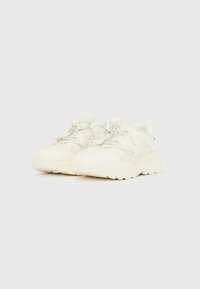 Pár törtfehér, masszív tornacipő texturált talppal és patentos rögzítéssel, egyszínű fehér háttéren.