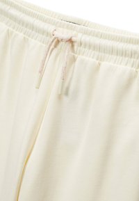 Pantalons de jogging couleur crème avec une taille élastique à cordon de serrage, présentant une texture de tissu douce et une finition élastique. Le cordon de serrage a un accent rose.