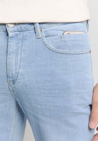 Lichtblauwe denim jeans met een klassieke vijf-pocket stijl, subtiele stiksels en een smartphone die gedeeltelijk zichtbaar is in de voorkennis.