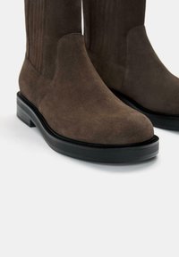 Botine din suede maro, cu vârful rotund, talpă groasă neagră și accente nervurate verticale pe tijă. Textură netedă.