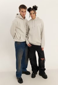 Dva mlada odrasla stojita drug ob drugem, oba oblečena v svetlo sive kapuce; eden v modrih kavbojkah, drugi v črnih raztrganih hlačah in črnih čevljih.