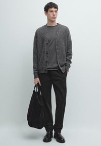 Grå texturerad kofta med knappar över en mörkgrå skjorta, svarta byxor och svarta läderskor, hållande en mörk tygväska.