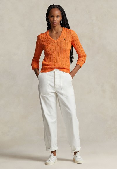 Pull en tricot torsadé orange avec un col en V ; associé à un pantalon blanc ample. Modèle portant des baskets blanches, mettant en avant un style vestimentaire décontracté.