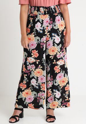 Czarne spodnie z szerokimi nogawkami z różowym, pomarańczowym i białym kwiatowym wzorem, noszone z czarnymi sandałami na obcasie i różowym krótkim topem.