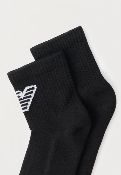 Ζευγάρι μαύρες ριγωτές κάλτσες μέχρι τον αστράγαλο με λευκό γεωμετρικό λογότυπο κοντά στη μανσέτα, τοποθετημένες επίπεδα σε λευκό φόντο.