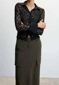 Chemise en dentelle noire à manches longues et col noir, associée à une jupe taille haute vert olive avec poches latérales et silhouette droite.