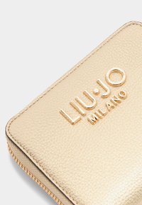 Zelta teksturēta maku ar reljefa "LIU-JO MILANO" logo, ar gludu rāvējslēdzēja aizdari un kontrastējošiem tumšiem malām.