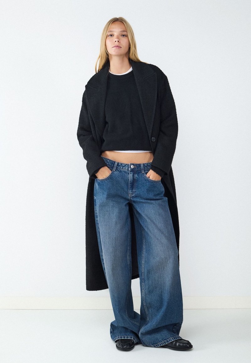 Woman wearing a black long coat, black cropped sweater, wide blue jeans, and black shoes standing with hands in pockets against a white wall.