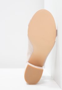 Sandale avec semelle lisse beige clair et petit talon bloc, positionnée verticalement contre un mur d'angle blanc.