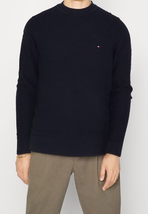 Homme portant un pull à manches longues noir texturé avec un petit logo sur la poitrine, accompagné d’un pantalon beige et d’un bracelet en or.